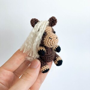 May include: A small, handmade crochet horse figurine. The horse features a brown body, tan face, and black hooves. It has a white, fluffy mane and dark brown ears. The miniature horse is held against a white background.