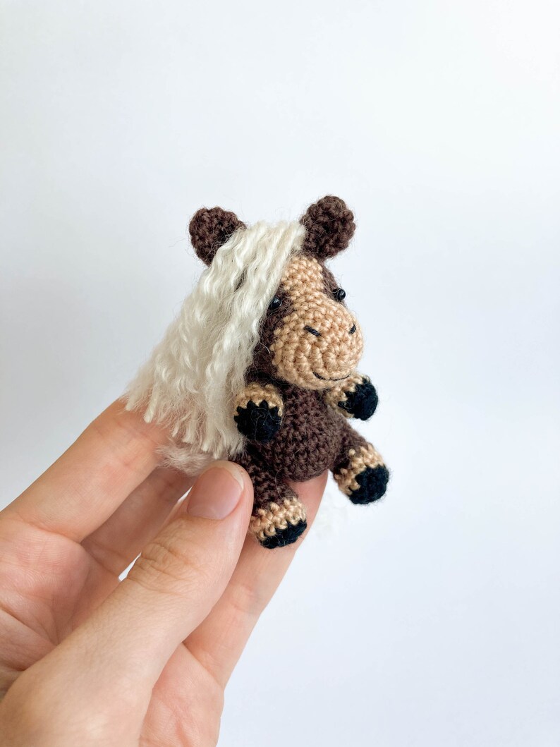 May include: A small, handmade crochet horse figurine. The horse features a brown body, tan face, and black hooves. It has a white, fluffy mane and dark brown ears. The miniature horse is held against a white background.