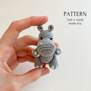 May include: A small, gray crocheted hippo toy with white accents on its feet and a rounded snout. The text "PATTERN *not a ready made toy" is visible in the upper right corner. The hippo is held in a hand against a white background.