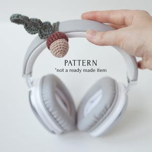 May include: A crocheted acorn charm with green leaves attached to a pair of gray headphones. The text "PATTERN *not a ready made item" is visible below the headphones.