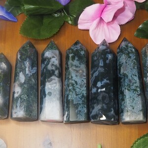 May include: Seven moss agate obelisks of varying heights, each with a pointed top. The stones display a mix of green, white, and translucent patterns. The obelisks are arranged on a wooden surface, with floral accents in the background.