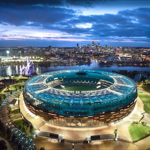 May include: An aerial view of a large, modern stadium with a blue and brown exterior, surrounded by green grass and a body of water. The stadium is lit up at night, and the city skyline can be seen in the background.