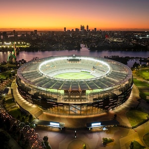 May include: An aerial view of a large, modern stadium with a green pitch and a retractable roof. The stadium is lit up at night, and the city skyline is visible in the background.