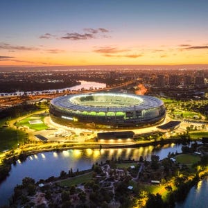 May include: An aerial view of a large stadium at dusk. The circular stadium has a green field and is surrounded by water and trees. The sky is a gradient of orange and blue.