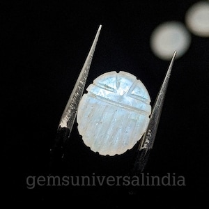 May include: A white moonstone cabochon carved with a geometric design. The stone is held in tweezers against a black background.