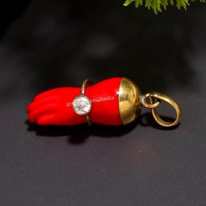 Puede incluir: Un colgante rojo en forma de mano con una tapa y un lazo dorados. Un anillo plateado con una gema transparente está en la mano. El colgante tiene el texto "geversalindia".