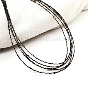 May include: A strand of black beads, made of small, rectangular shapes, is draped over a white fabric surface. The beads are strung together to form a necklace.