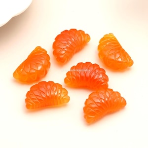 May include: A collection of orange-hued, carved gemstone segments, designed to resemble mandarin orange slices. The translucent stones are displayed on a white surface, highlighting their detailed, textured patterns. These segments are likely intended for jewellery or decorative use.