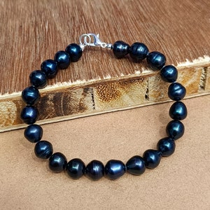 Puede incluir: Un brazalete de perlas negras con un cierre plateado. Las perlas son redondas y tienen un ligero brillo iridiscente.