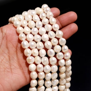 Pode incluir: Um fio de pérolas de água doce brancas com uma forma orgânica irregular e um acabamento lustroso e iridescente. As pérolas estão enfiadas numa linha contínua, exibindo a sua textura natural, tamanhos variados e superfícies lisas e reflexivas.