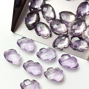 May include: A collection of purple amethyst gemstone beads in a faceted, flower-shaped design. The beads are arranged on a silver tray and a white surface.