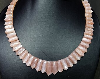 May include: A necklace made of light pink sunstone beads. The beads are faceted and have a pointed shape. The necklace is strung on a gold cord with red tassels at the ends.