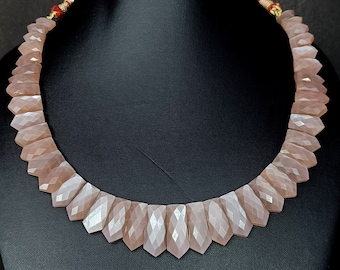 May include: A necklace made of light pink faceted gemstone beads. The beads are arranged in a pointed, chevron pattern. The necklace is displayed on a black mannequin.