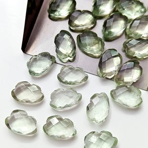 May include: A selection of pale green, faceted gemstones in diverse shapes, including teardrops and cloud-like forms. The gems are presented on a silver tray, highlighting their clarity and light-reflecting properties. The overall aesthetic is one of delicate beauty and natural elegance.