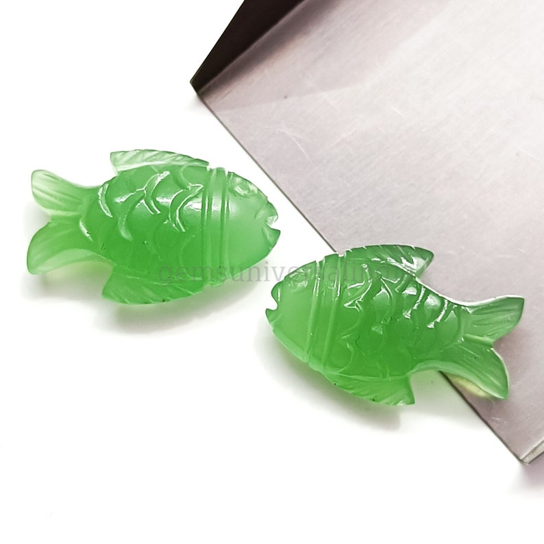 K&ouml;nnte beinhalten: Zwei gr&uuml;ne Jade-Fisch-Anh&auml;nger mit detaillierten Schnitzereien. Die Fische sind in entgegengesetzte Richtungen gerichtet und haben eine glatte, polierte Oberfl&auml;che.