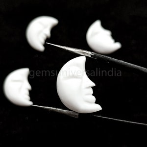 Puede incluir: Cuatro cabujones blancos en forma de media luna, uno sostenido por unas pinzas, con una cara tallada en la luna. Los cabujones están hechos de concha o hueso blanco.