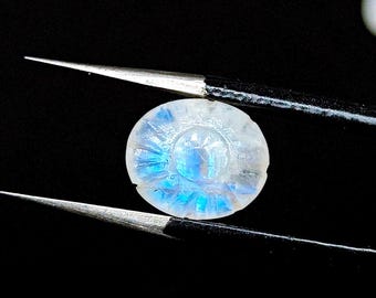 Può includere: Un cabochon di pietra di luna bianca con un'iridescenza blu. La pietra è rotonda e presenta un design intagliato sulla superficie.
