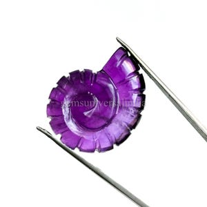 May include: A carved, purple gemstone in a spiral seashell design is held by silver tweezers against a white background. The gemstone has a polished, glossy finish and is intricately detailed.