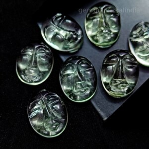 May include: Seven green gemstone cabochons carved with faces. The cabochons are oval shaped and have a smooth, polished finish.