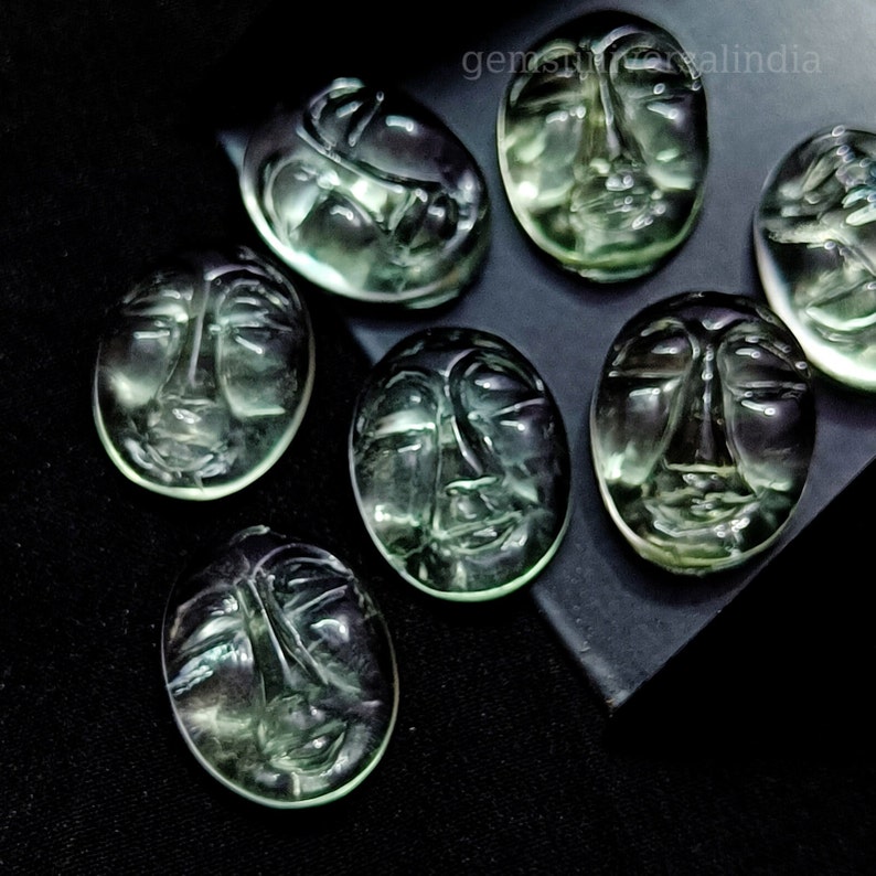 May include: Seven green gemstone cabochons carved with faces. The cabochons are oval shaped and have a smooth, polished finish.