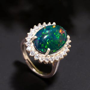 May include: A gold-toned ring featuring a large, oval-shaped black opal with vibrant green and red flashes. The opal is surrounded by a halo of small, clear gemstones, creating a sparkling effect. The ring is set against a dark background.