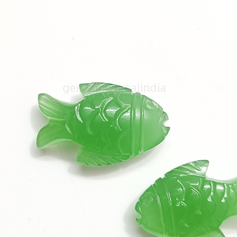 K&ouml;nnte beinhalten: Zwei gr&uuml;ne Jade-Fisch-Anh&auml;nger mit detaillierten Schnitzereien. Die Fische sind in entgegengesetzte Richtungen ausgerichtet und haben eine glatte, polierte Oberfl&auml;che.