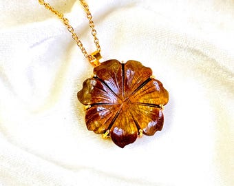 May include: A gold-toned chain necklace featuring a pendant carved from brown tiger's eye gemstone into the shape of a six-petaled flower. The stone displays natural chatoyant bands and is secured by small gold-toned prongs at the edges.