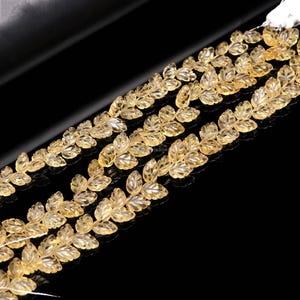 May include: Close-up of several strands of yellow leaf-shaped beads. The beads are translucent and have a detailed leaf design. The background is black, highlighting the beads' color and shape. These beads are likely for jewelry making.