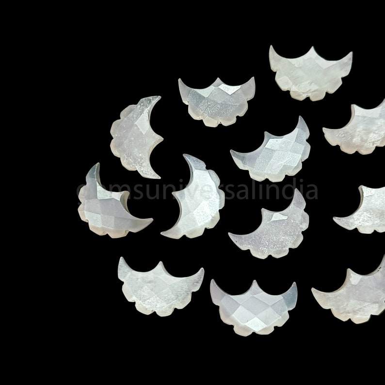 May include: A collection of 12 white, crescent moon shaped, mother of pearl gemstone cabochons. The cabochons are faceted and have a slightly iridescent sheen.