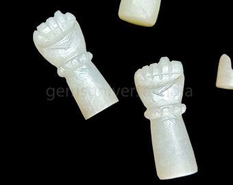 Könnte beinhalten: Zwei weiße Perlmutt-Anhänger in Form einer Faust. Die Anhänger sind mit einem erhabenen Design und einem Band um das Handgelenk verziert.