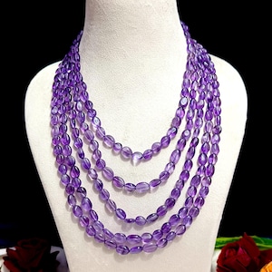 May include: A multi-strand necklace featuring oval-shaped amethyst beads in varying lengths. The beads are a deep purple hue, strung together to create a layered effect. The necklace is displayed on a white mannequin bust against a dark background.