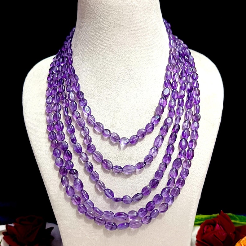 May include: A multi-strand necklace featuring oval-shaped amethyst beads in varying lengths. The beads are a deep purple hue, strung together to create a layered effect. The necklace is displayed on a white mannequin bust against a dark background.