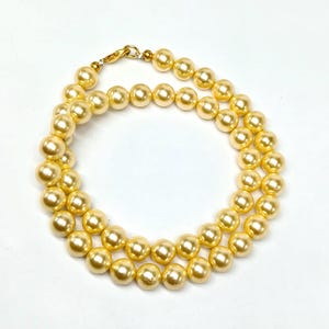 Puede incluir: Un collar de perlas doradas con cierre de broche. Las perlas son redondas y tienen un acabado liso y brillante.