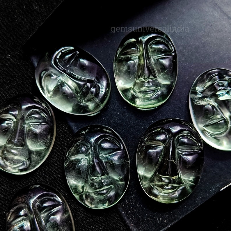 May include: Eight green face carvings, each with a unique expression, are displayed on a black surface. The carvings are made of a translucent material, possibly green aventurine or green fluorite.