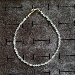 Könnte beinhalten: Eine zarte silberne Kettenkette mit klaren Kristallperlen. Die Kette liegt auf einer grauen, strukturierten Oberfläche.