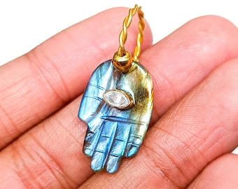 Könnte beinhalten: Goldener Ohrring mit einem handförmigen Anhänger. Der Anhänger ist blau und goldfarben mit einem geschnitzten Design und einem klaren, oval geformten Stein. Der Ohrring hat einen gedrehten Goldreifen.