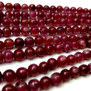 May include: A strand of deep red ruby beads, each bead is round and smooth with a slight shimmer. The beads are arranged in a loose pile, showcasing their color and texture.