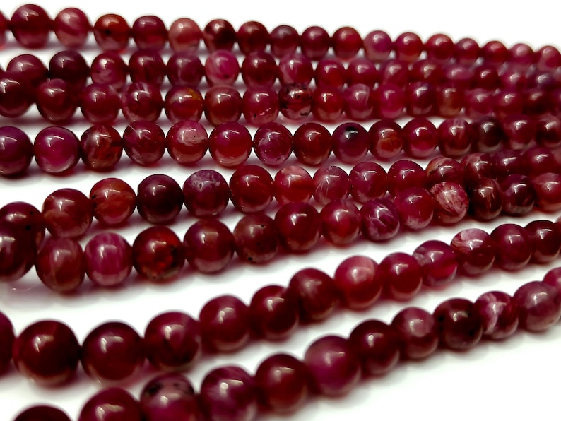May include: A strand of deep red ruby beads, each bead is round and smooth with a slight shimmer. The beads are arranged in a loose pile, showcasing their color and texture.