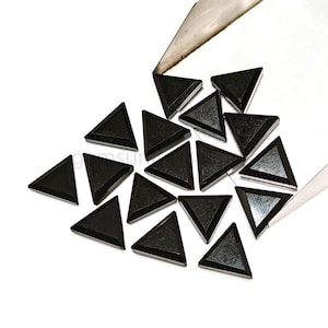 May include: A collection of black triangle-shaped glass gems. The gems are scattered on a white surface and are being poured from a metal container.