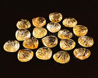 May include: A collection of 16 carved, translucent, yellow-gold citrine stones in a heart shape. The stones have a striated pattern and are arranged on a black surface.