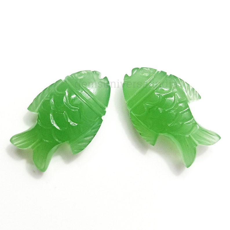 K&ouml;nnte beinhalten: Zwei gr&uuml;ne Jade-Fisch-Anh&auml;nger mit aufwendigen Schnitzereien. Die Fische sind in entgegengesetzte Richtungen gerichtet und haben eine glatte, polierte Oberfl&auml;che.