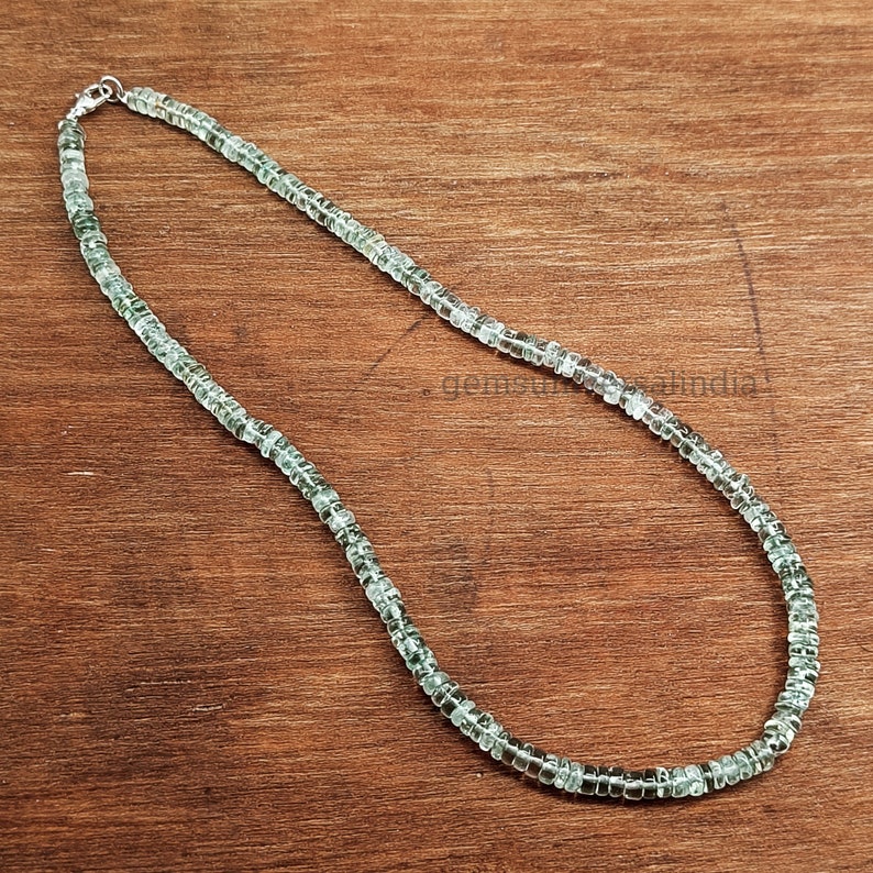 May include: A silver chain necklace with light green square beads. The necklace is on a wooden surface.