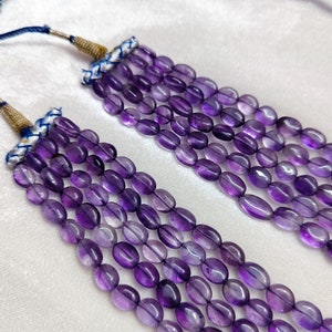 May include: Close-up of multiple strands of amethyst gemstone beads. The beads are oval-shaped and a deep purple color. The strands are attached to a gold-colored cord with blue and white accents. The background is a soft white fabric.