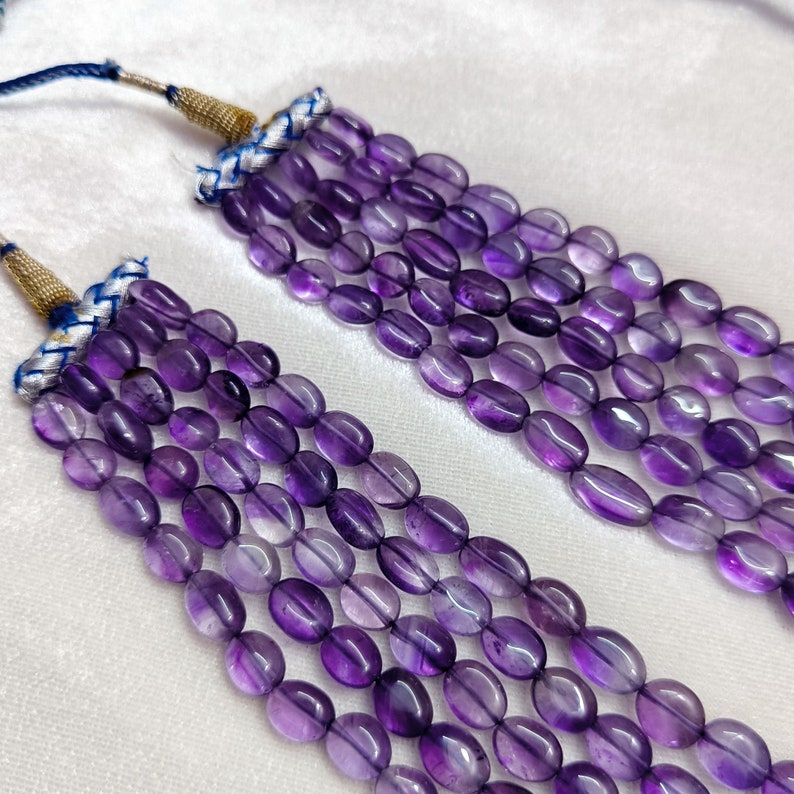 May include: Close-up of multiple strands of amethyst gemstone beads. The beads are oval-shaped and a deep purple color. The strands are attached to a gold-colored cord with blue and white accents. The background is a soft white fabric.