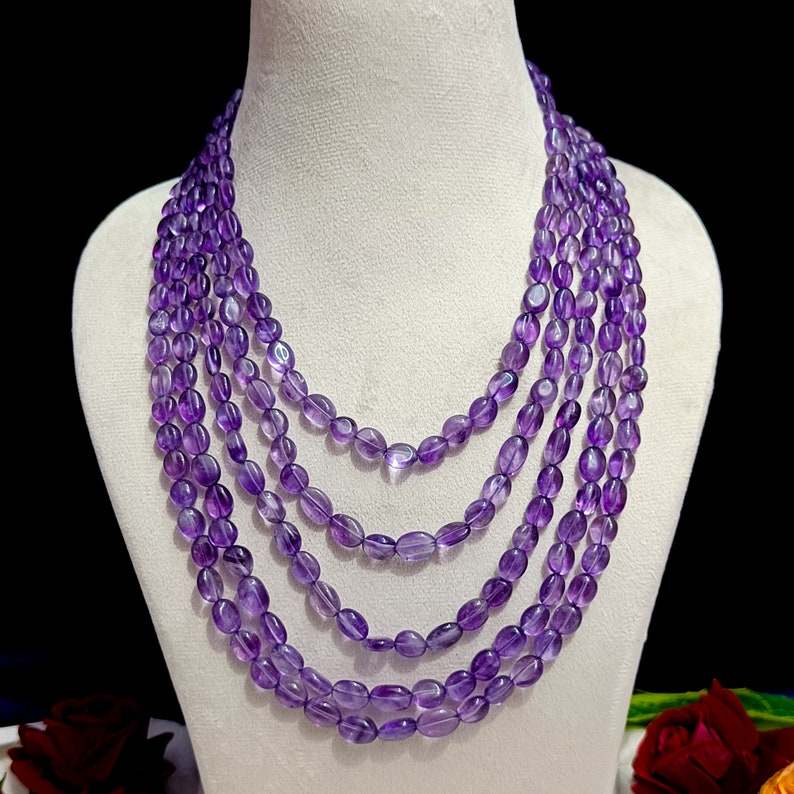 May include: A multi-strand necklace composed of oval-shaped, deep purple amethyst beads. The necklace is displayed on a white bust form against a dark background. The beads are strung together in multiple layers, creating a layered effect.