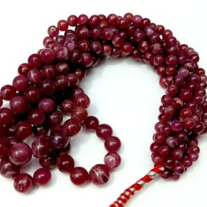 May include: A strand of deep red gemstone beads with a red and silver cord. The beads are round and have a smooth, polished finish.