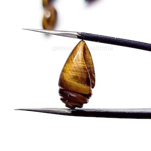 May include: A brown and gold tiger's eye gemstone carved into a teardrop shape. The gemstone is held by a pair of tweezers. The text "gemsunsalindia" is visible in the background.