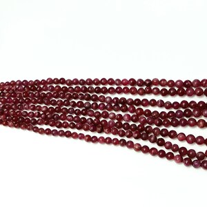 May include: A strand of deep red ruby beads, each bead is round and smooth. The beads are strung together on a clear thread.