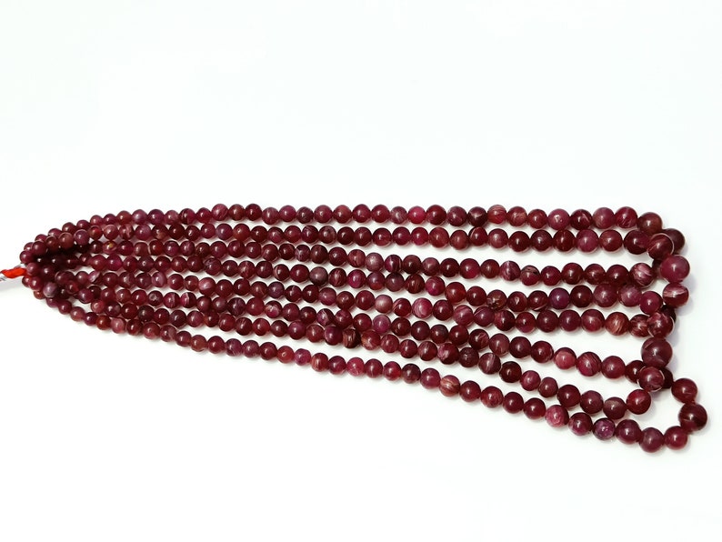 May include: A strand of deep red ruby beads, each bead is round and smooth. The beads are strung together on a clear thread.
