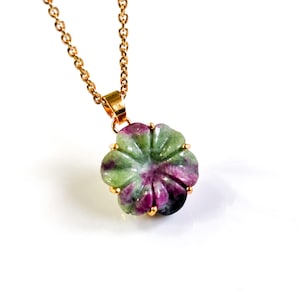 Può includere: Una collana con catena color oro con un ciondolo a forma di fiore. Il ciondolo è scolpito in una pietra con sfumature verdi e magenta. Il fiore ha sei petali ed è incastonato in una lunetta color oro.